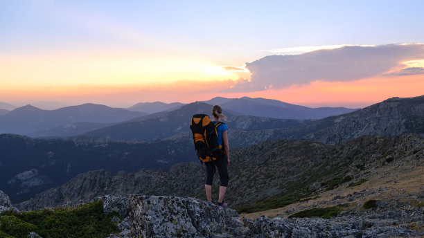 一名女子徒步旅行者站在以山脉为背景的一块大岩石上观看日落。图片下载