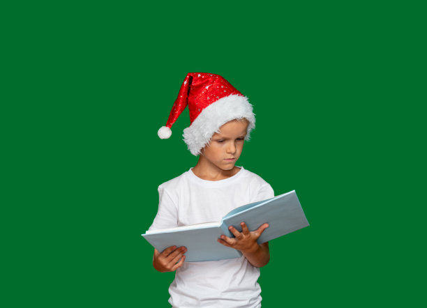 戴着圣诞老人帽子的男孩正在思考，看着一本打开的书。绿色背景与文本空间。有选择性的重点。关于儿童、节日、圣诞节的文章和广告图片。图片下载