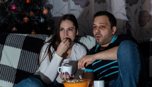 年轻的女人和男人拿着马克杯，一边看电视一边吃爆米花。特写镜头。有选择性的重点。关于电影、爱好、休闲的文章图片。图片下载