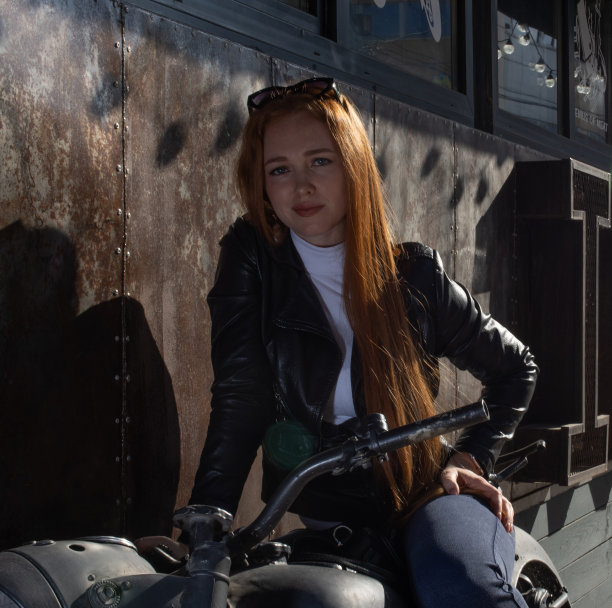 一个长着红色长发的漂亮女孩，穿着皮夹克，在摩托车上摆姿势。图片下载