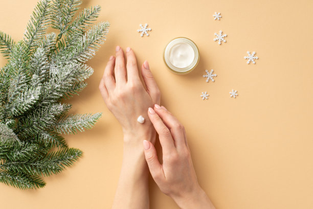 冬季护肤理念。第一人称俯视图照片，女子的手应用霜上她的手从小罐子冷杉树枝在白霜和雪花孤立的粉彩米色背景图片下载