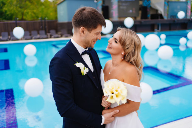 新娘和新郎穿着经典的衣服，站在装饰着白色气球的泳池边。图片下载