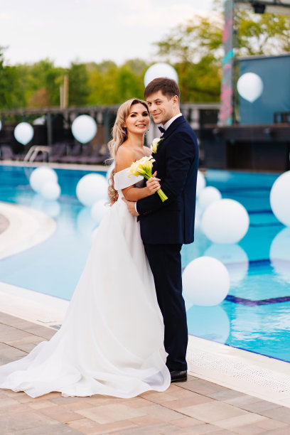 新娘和新郎穿着经典的衣服，站在装饰着白色气球的泳池边。图片下载