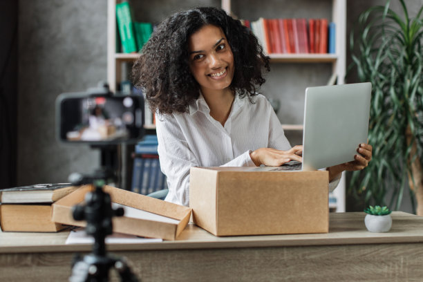 多元文化的妇女用手机摄像头录制视频，同时用新的无线笔记本电脑打开包装盒。图片下载