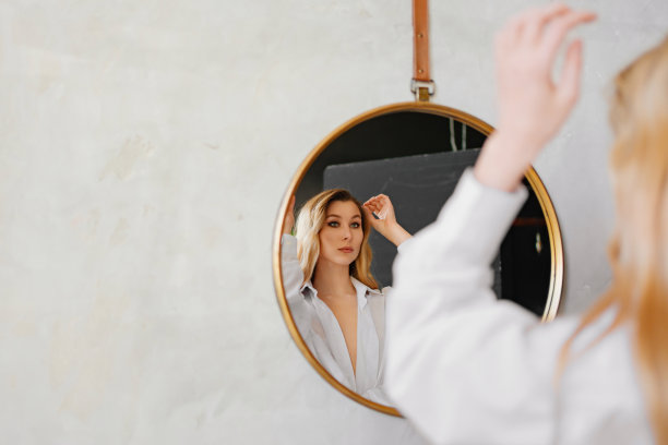 一个穿着白衬衫的金发美女正看着圆镜中的自己。图片下载