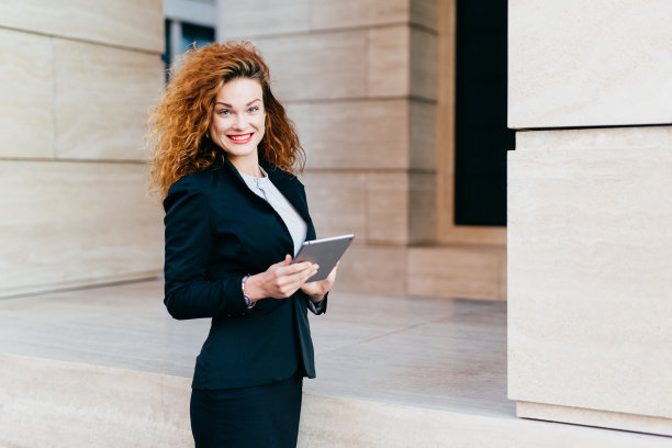 人、业务、创新和技术理念。快乐的卷发美女涂着红色的嘴唇，穿着正式的衣服，拿着现代的平板电脑，对着她的办公大楼摆姿势图片下载
