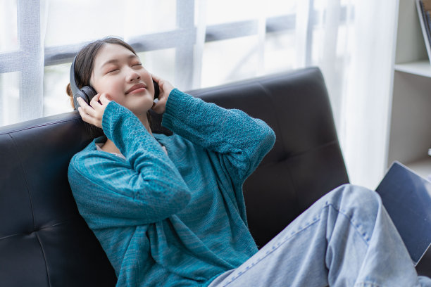 年轻的亚洲女人在家里客厅的沙发上听音乐。快乐的女人在度假时使用智能手机图片下载
