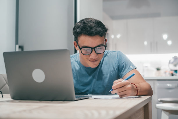 一个年轻的青少年学习和工作，在家里的桌子上使用笔记本电脑，微笑着做作业。图片下载