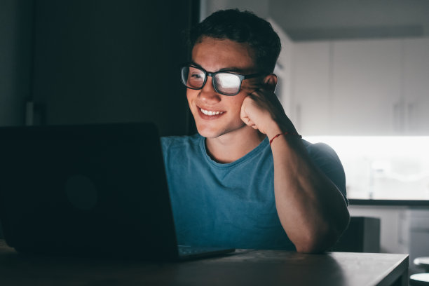 一个年轻的青少年学习和工作，在家里的桌子上使用笔记本电脑，微笑着做作业。屏幕的光线照在脸上。图片下载