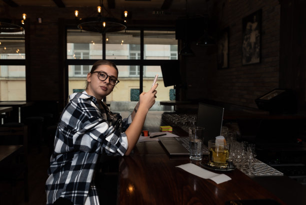 年轻的自由职业商业女性坐在咖啡厅的吧台上，在她的笔记本电脑上工作，休息一下，在她的智能手机上玩社交媒体。工作之余的专业商务人士。图片下载