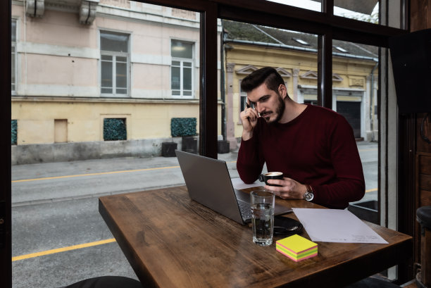 年轻的商人坐在咖啡馆里，在午休时间喝咖啡，在笔记本电脑上工作。为外国公司从事新项目的自由职业者。图片下载