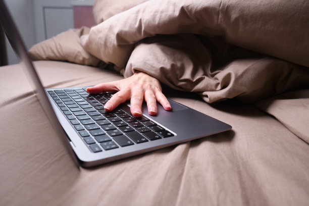 一名年轻女子正裹着毯子躺在床上用笔记本电脑工作。在家工作，自由职业者。关注手图片下载