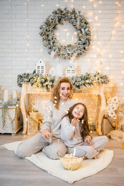 年轻女子带着7岁的小女孩穿着睡衣在圣诞装饰的家里看电影和吃爆米花图片下载