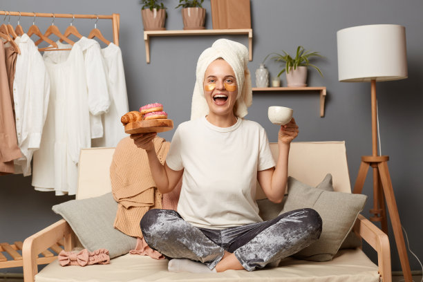 兴奋快乐的女人在她头上的毛巾和眼罩的照片，坐在咳嗽与架子上的衣服背景，有美味的小吃和喝咖啡，享受咖啡。图片下载