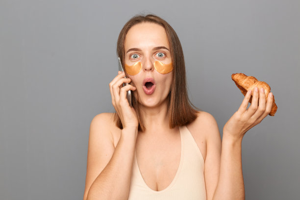 室内照片，震惊的女子打电话，在眼睛下面涂上美容眼罩，在灰色背景下，在室内孤立地摆姿势，拿着羊角面包，张着嘴。图片下载