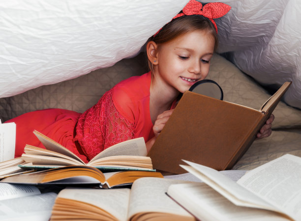可爱的6-7岁女孩读书。小女孩在家里的床上穿着红裙子，盖着毯子，前景上有许多书。用放大镜搜索书中的信息图片下载