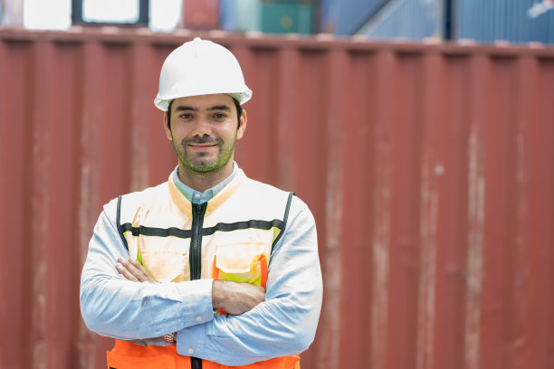 快乐的男性集装箱堆场工人在商业码头工地工作的肖像。男性领班或主管站在集装箱堆场前控制装载集装箱的箱子图片下载