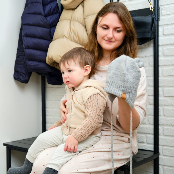 母亲给坐在家里走廊上的蹒跚学步的婴儿戴上一顶温暖的帽子。在寒冷的天气里，妈妈给孩子穿保暖的衣服。一岁零三个月的孩子图片下载