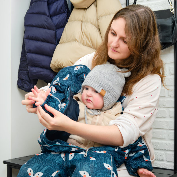 母亲给坐在家里走廊上的蹒跚学步的婴儿穿上了一件蓝色的工作服。在寒冷的天气里，妈妈给孩子穿上保暖的衣服。一岁零三个月的孩子图片下载