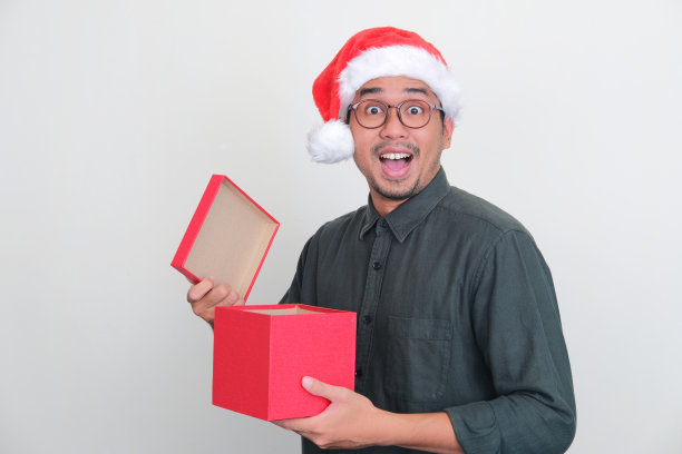 亚洲男子戴着圣诞帽，打开一盒礼物时露出惊叹的表情图片下载
