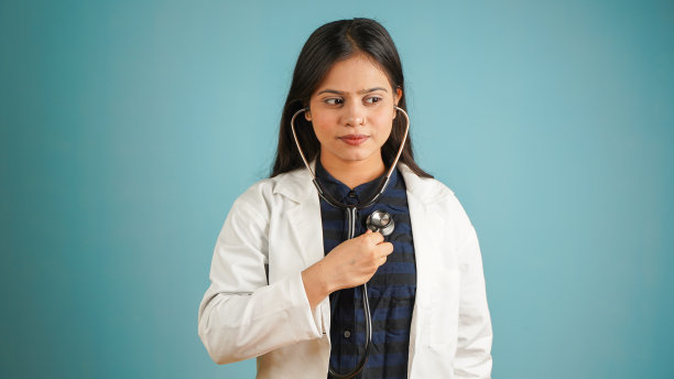一个年轻的女医生听她的心跳，亚裔印度女医生在围裙和听诊器隔离在蓝色的工作室背景图片下载