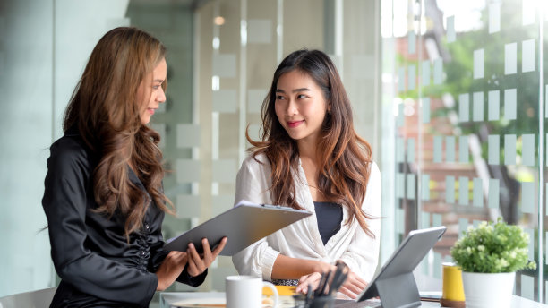 两个年轻漂亮的亚洲女商人坐在办公桌前，拿着笔记本电脑一起做文书工作，讨论项目财务报告。公司业务协作概念。图片下载
