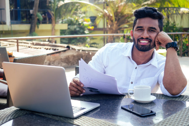 英俊而成功的印度男人穿着时尚得体的自由职业者在海滩上工作笔记本电脑，自由职业者和远程工作。商人学生在印度洋岸边的夏日咖啡馆里喝着茶图片下载