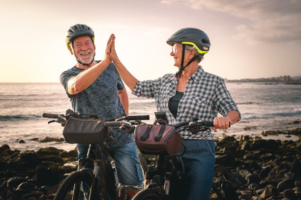 击掌吧。日落时分，一对兴高采烈的老夫妇骑着电动自行车在卵石海滩上散步。真实的老人退休生活和可持续的出行理念图片下载