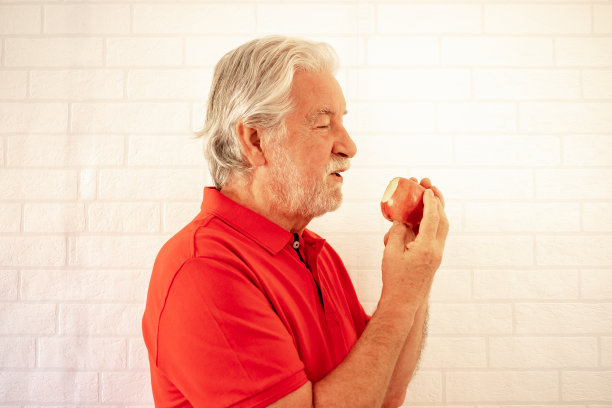 一位穿着红色polo衫的老人微笑着在白色背景下咬着苹果，这是健康饮食的概念图片下载