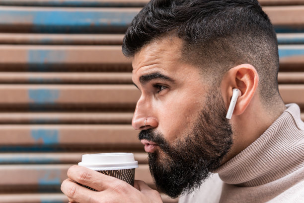 特写照片中，一名留着胡子、穿着高领毛衣的年轻男子一边喝着一次性咖啡，一边戴着无线耳机。图片下载