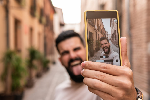 一个留着胡子、穿着高领毛衣的年轻人在他所在城市最著名的街道之一吐着舌头自拍。图片下载