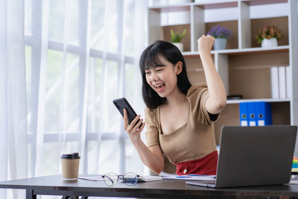 兴奋的亚洲女商人，穿着西装，微笑着，尖叫着，紧握拳头，在办公室的电脑屏幕和智能手机前庆祝胜利图片下载