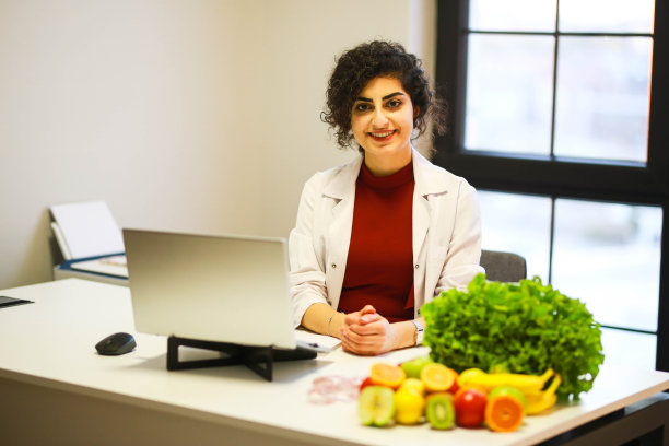 微笑的黑发女性营养师在她的办公室写饮食计划，显示健康的蔬菜和水果。健康和饮食概念。生活方式。图片下载