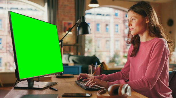 年轻漂亮的女性在家用台式电脑上工作，绿色屏幕模拟显示。查看社交媒体，浏览互联网。阁楼公寓，从大窗户可以看到城市景观。图片下载