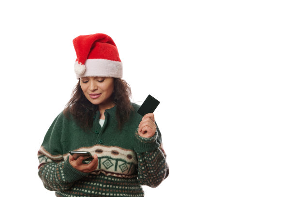 快乐的女顾客戴着圣诞帽和圣诞图案的毛衣，使用手机应用程序，智能手机和信用卡，网上购物图片下载
