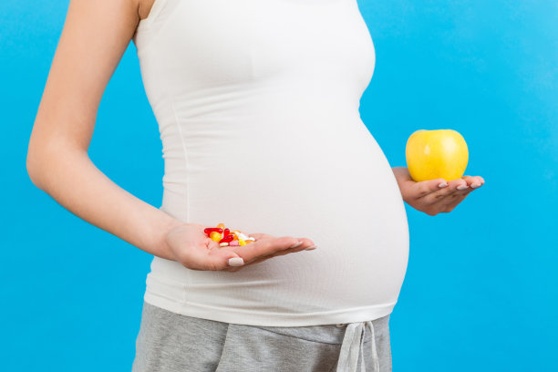 特写孕妇在苹果和一堆药丸之间做出选择，彩色背景与复制空间。怀孕期间服用维生素的概念图片下载
