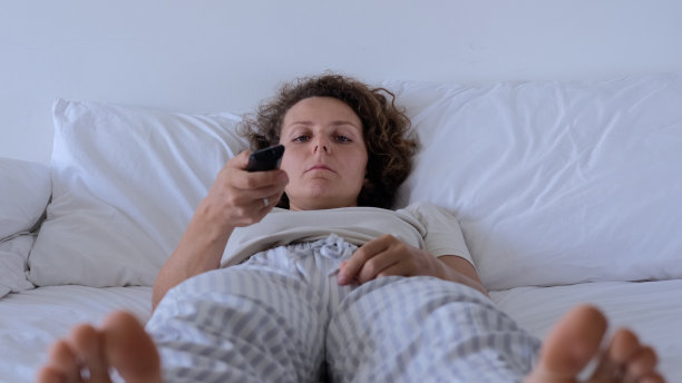 一个年轻女子躺在床上，手里拿着电视遥控器，无聊地切换着频道。一个抑郁的女人独自躺在床上看电视。在卧室的床上休息。图片下载