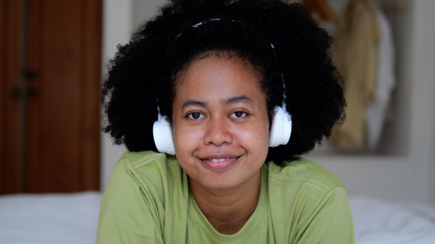一个年轻的爆炸头女子，黑色美丽的卷发，有趣，冷静和友好的心情，她的空闲时间躺在床上，听音乐，戴着耳机在一个舒适的房间。图片下载