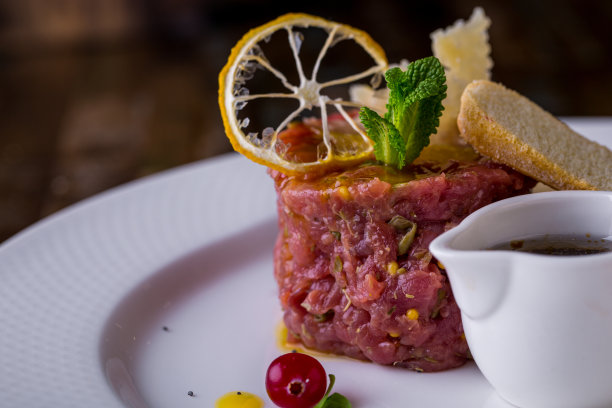 小牛肉塔塔配第戎芥末和芒果酱。上面是一片柠檬片、一片薄荷叶、一片帕尔玛干酪片和白面包吐司。旁边是一个盛满了腌制焦糖洋葱的肉卤盘。食物在一个白色的盘子里。图片下载