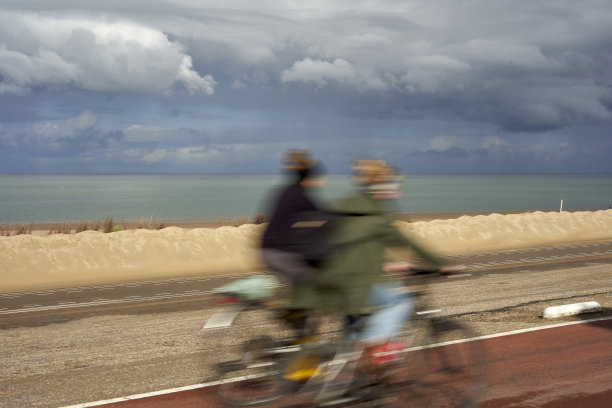 在荷兰，两名女子骑自行车运动。年轻女子骑着自行车。在夏天的雨天。多云天空下的北海。荷兰，泽兰，布鲁沃斯丹。图片下载