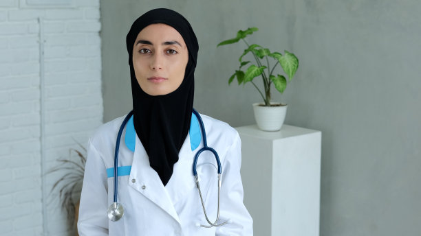 一名阿拉伯妇女站在办公室里，戴着黑色头巾，一件有蓝色插孔的医疗服，头巾上挂着一个医疗设备。冷静自信的伊斯兰女医生。图片下载