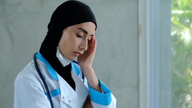 一名穆斯林女医生，戴着黑色头巾，穿着白色的蓝色插孔医疗服，从脸上摘下医用口罩，按摩太阳穴。阿拉伯女医生累了。图片下载