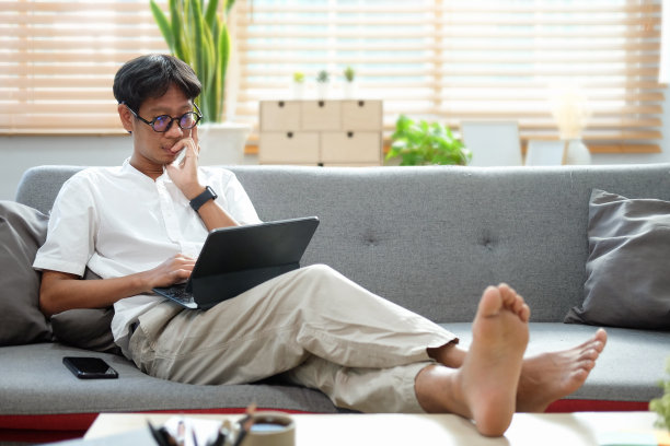 随意的亚洲男人坐在沙发上，用着平板电脑。图片下载