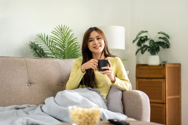 年轻的亚洲女子正在喝咖啡，躺在舒适的大沙发上看电视电影图片下载