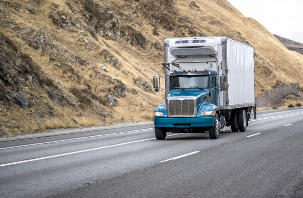 紧凑的日间驾驶室大钻机半卡车和冷藏箱拖车运输货物行驶在宽阔的多线公路上，两侧有岩石山图片下载