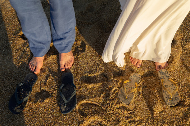 婚纱照片。新郎和新娘并肩站在沙滩上。附近有两双凉鞋。金色的光芒穿透。图片下载