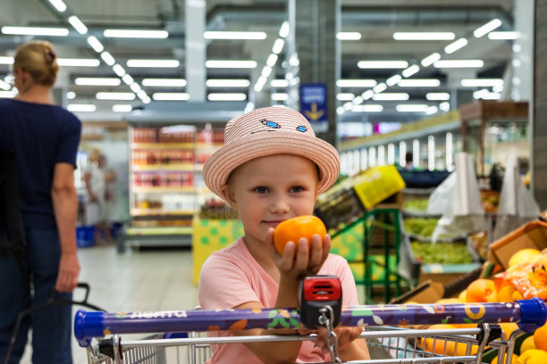 有趣的小女孩与普通话坐在购物车在食品水果店，高兴地看着相机。可爱的孩子购物和购买零售新鲜水果。儿童健康食品概念。复制文本空格图片下载