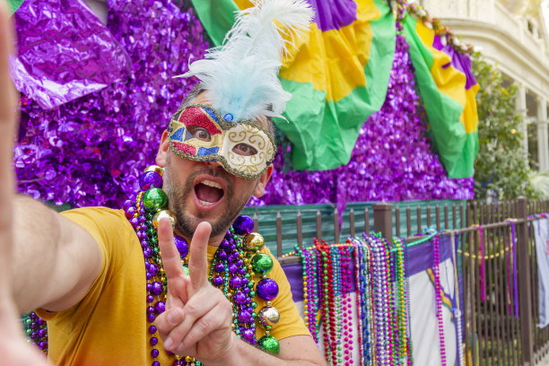 一名年轻的拉丁男性游客，戴着面具，穿着服装，戴着项链，在新奥尔良的街道上庆祝狂欢节，同时用手机自拍。图片下载