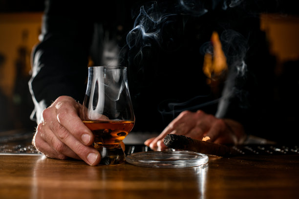男性手拿酒杯和烟灰缸，烟灰缸附近有一支正在吸烟的古巴雪茄图片下载