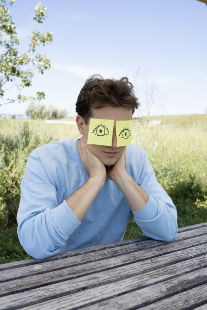 一个穿着蓝色毛衣的年轻人坐在公园里，他的头上有两张黄色的纸币，上面画着眼睛图片下载